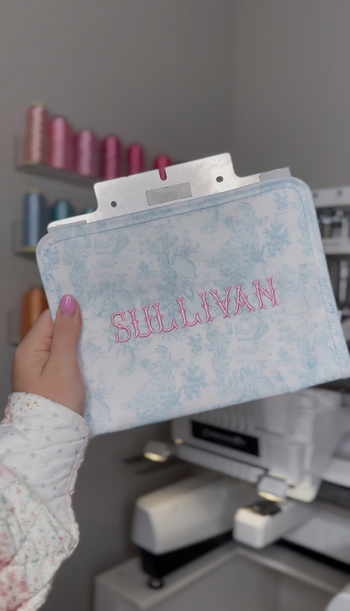 Light blue bunny toile TRVL pouch embroidered with the name 'Sullivan' in pink, shown on an embroidery machine
