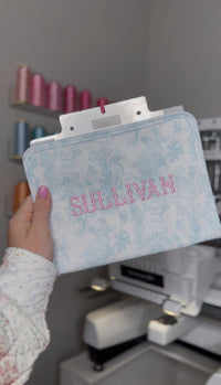Light blue bunny toile TRVL pouch embroidered with the name 'Sullivan' in pink, shown on an embroidery machine