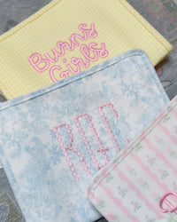 Blue bunny toile TRVL pouch with pink beaded monogram, next to a yellow gingham pouch embroidered 'Burns Girls'