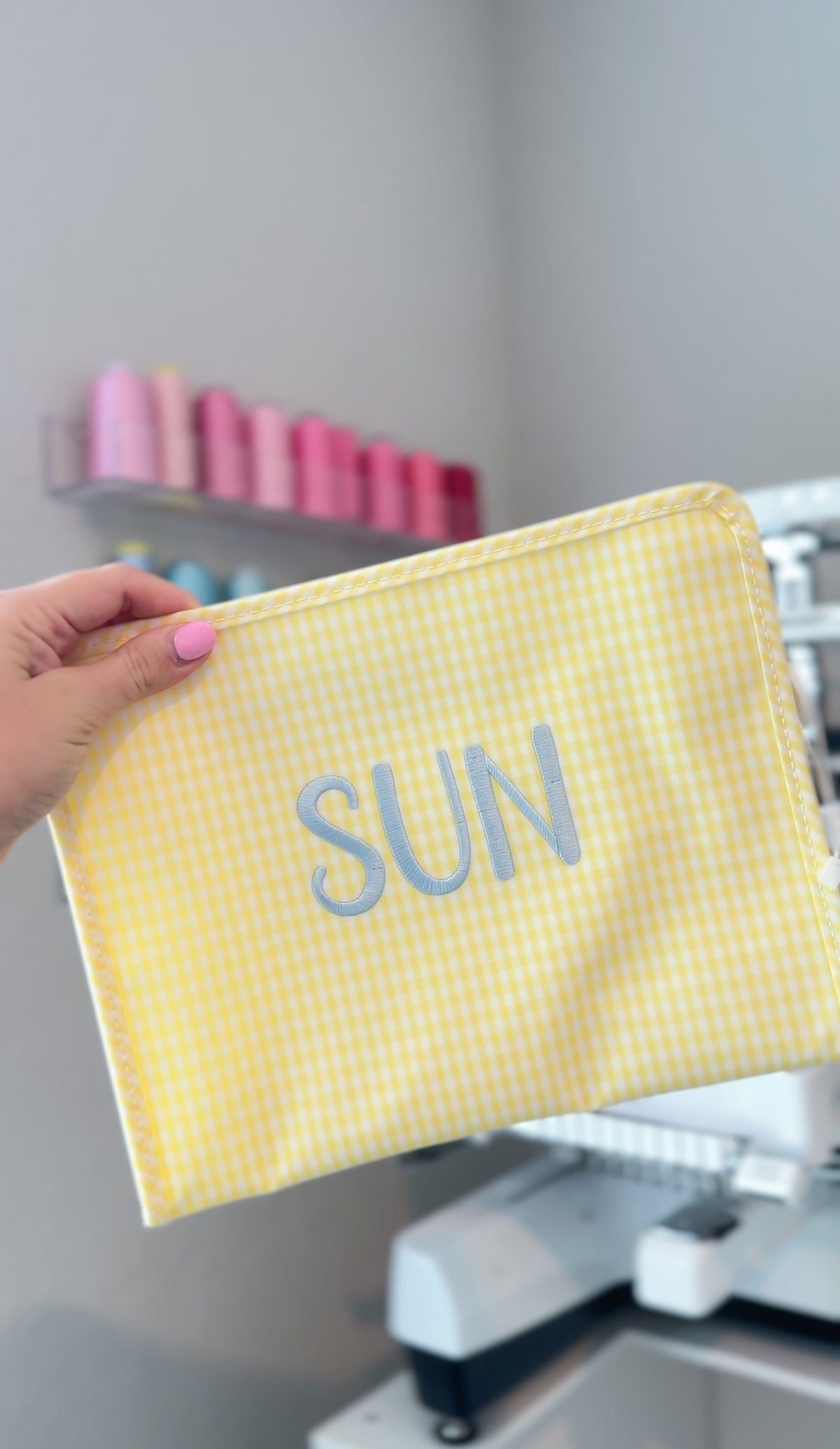 Yellow gingham TRVL Roadie pouch embroidered with 'SUN' in silver block lettering, held up in a monogramming studio