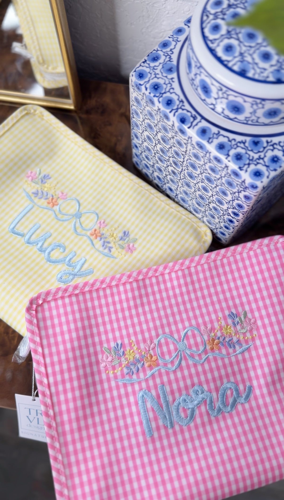 Yellow gingham TRVL Roadie pouch embroidered with the name 'Lucy' in a floral bow design, next to a pink gingham pouch embroidered 'Nora'