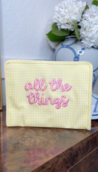 Yellow gingham TRVL pouch embroidered with 'all the things' in pink script, displayed near a blue and white ginger jar