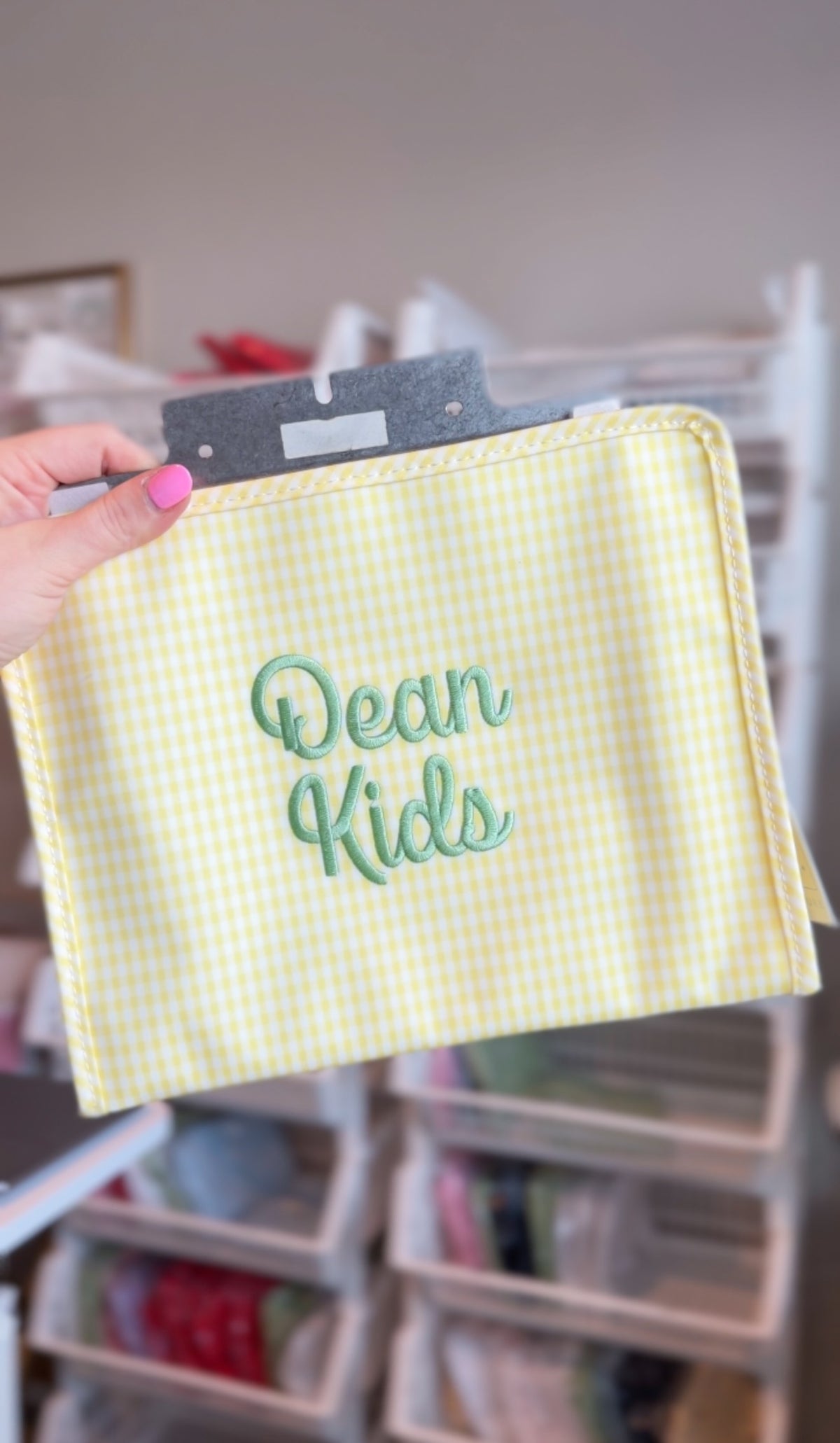 Yellow gingham TRVL pouch embroidered with 'Dean Kids' in sage green script, held up in a storage room