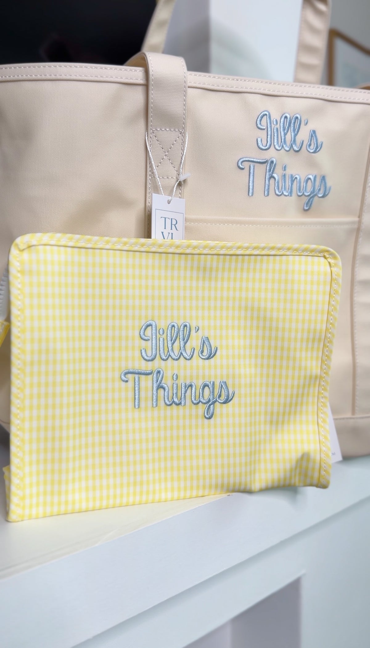 Yellow gingham TRVL pouch embroidered with 'Jill's Things' in light blue script, shown in front of a matching tan tote with the same embroidery