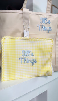 Yellow gingham TRVL pouch embroidered with 'Jill's Things' in light blue script, shown in front of a matching tan tote with the same embroidery