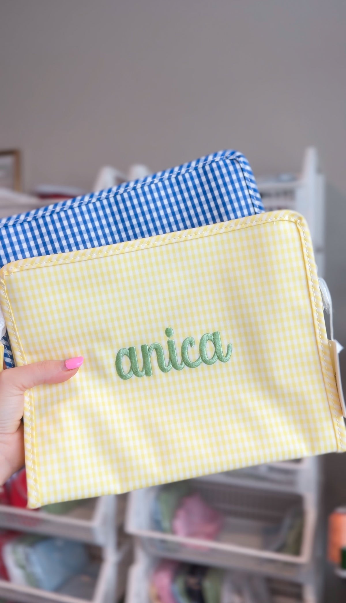 Yellow gingham TRVL pouch embroidered with the name 'anica' in sage green script, layered in front of a blue gingham pouch