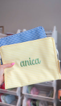 Yellow gingham TRVL pouch embroidered with the name 'anica' in sage green script, layered in front of a blue gingham pouch