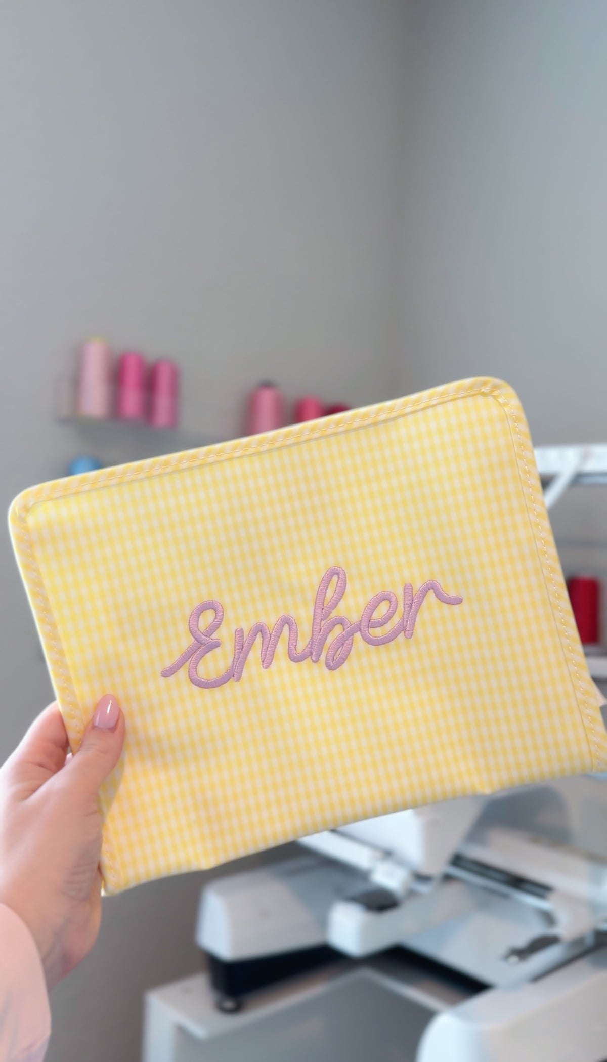 Yellow gingham TRVL Roadie pouch embroidered with the name 'Ember' in lilac script, held up in a monogramming studio