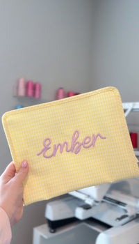 Yellow gingham TRVL Roadie pouch embroidered with the name 'Ember' in lilac script, held up in a monogramming studio