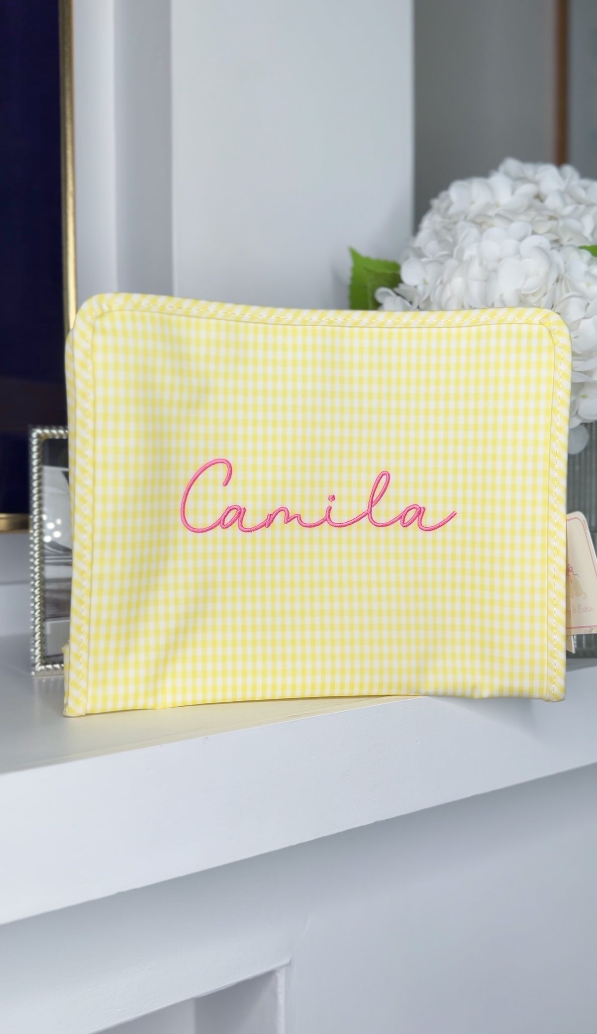 Yellow gingham TRVL Roadie pouch embroidered with the name 'Camila' in pink script, displayed on a mantel with hydrangeas