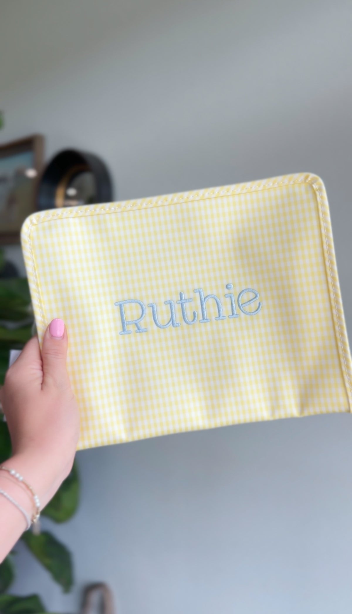 Yellow gingham TRVL Roadie pouch embroidered with the name 'Ruthie' in light blue block lettering, held up indoors