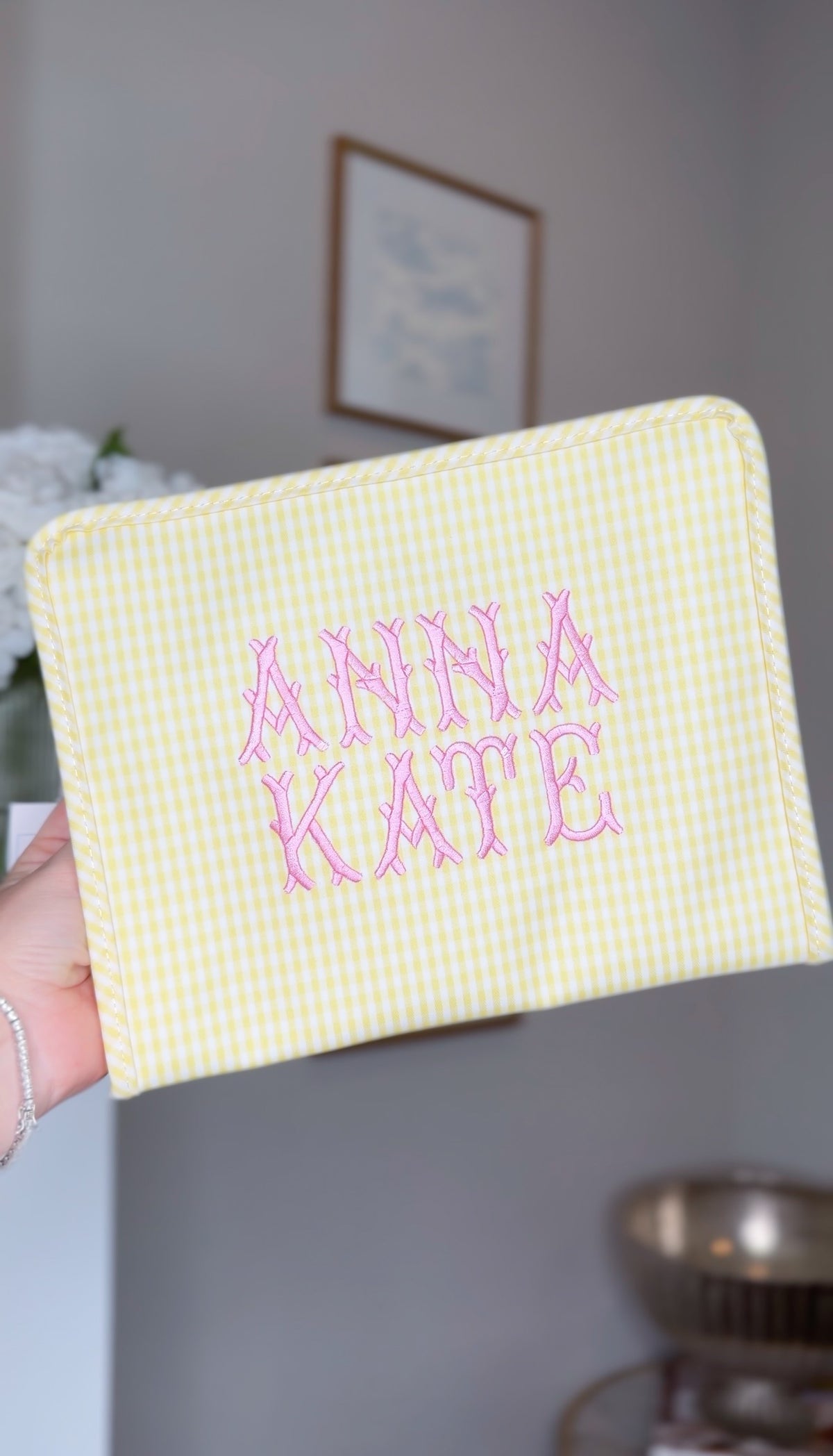 Yellow gingham TRVL Roadie pouch embroidered with 'Anna Kate' in pink block lettering, held up indoors