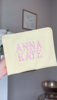 Yellow gingham TRVL Roadie pouch embroidered with 'Anna Kate' in pink block lettering, held up indoors