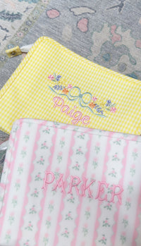 Yellow gingham TRVL Roadie pouch embroidered with a floral bow design and the name 'Paige', shown next to a pink floral striped pouch embroidered 'Parker'