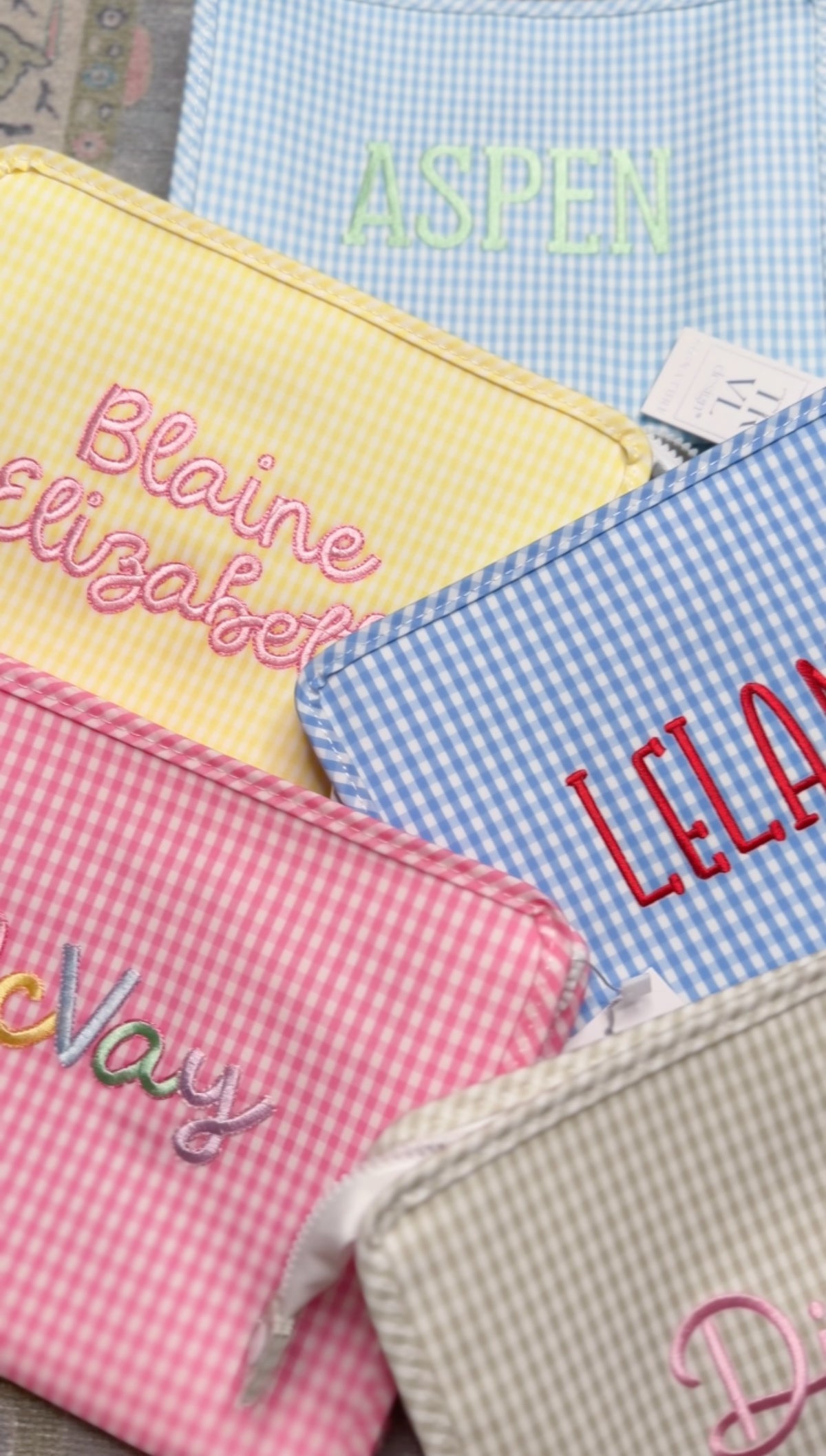 Assortment of gingham TRVL Roadie pouches in yellow, pink, and blue embroidered with names including 'Aspen', 'Blaine Elizabeth', 'Leland', and 'McVay'
