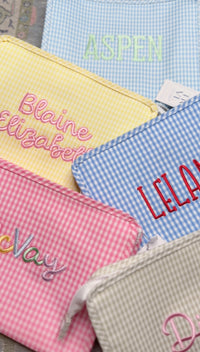 Assortment of gingham TRVL Roadie pouches in yellow, pink, and blue embroidered with names including 'Aspen', 'Blaine Elizabeth', 'Leland', and 'McVay'