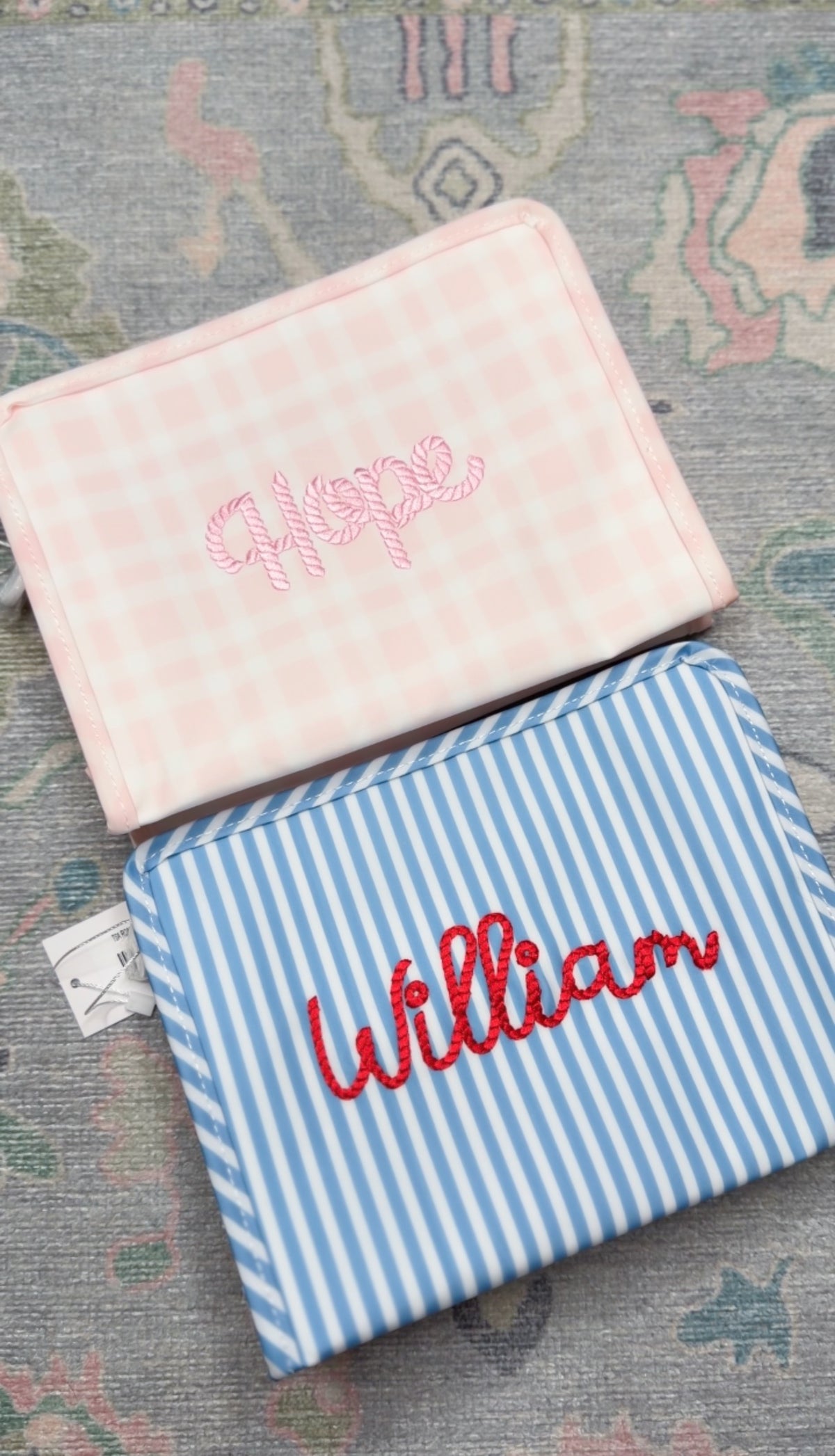 Blue chambray striped TRVL pouch embroidered 'William' in red, next to a pink gingham pouch embroidered 'Hope'