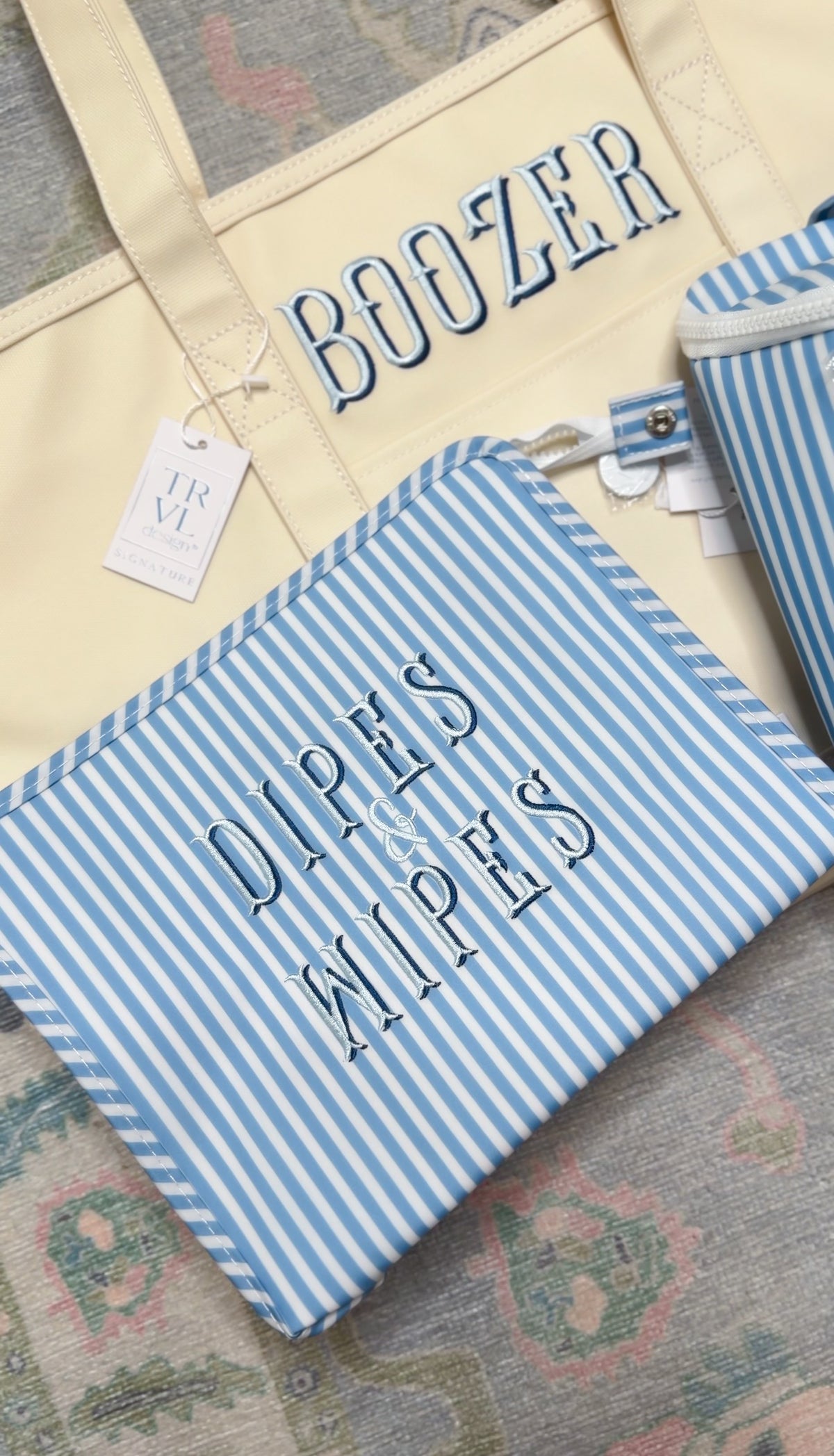 Blue chambray striped TRVL pouch embroidered 'Dipes & Wipes', next to a yellow tote embroidered 'Boozer'