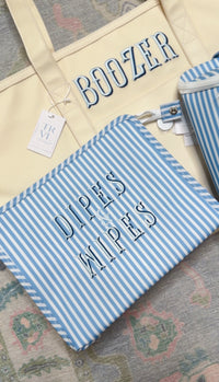 Blue chambray striped TRVL pouch embroidered 'Dipes & Wipes', next to a yellow tote embroidered 'Boozer'