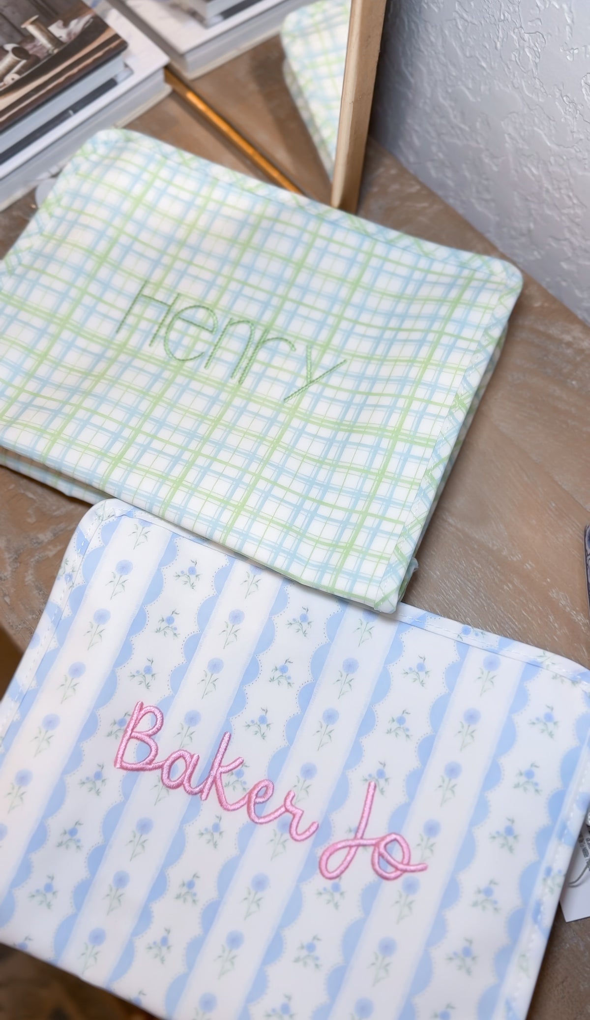 Green plaid TRVL pouch embroidered 'Henry', next to a blue floral pouch embroidered 'Baker Jo'