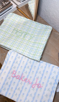 Green plaid TRVL pouch embroidered 'Henry', next to a blue floral pouch embroidered 'Baker Jo'