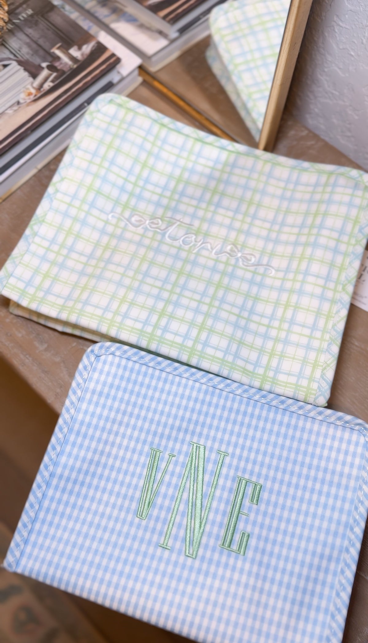 Green plaid TRVL pouch embroidered with a name in white script, next to a blue gingham pouch with the green 'VNE' monogram