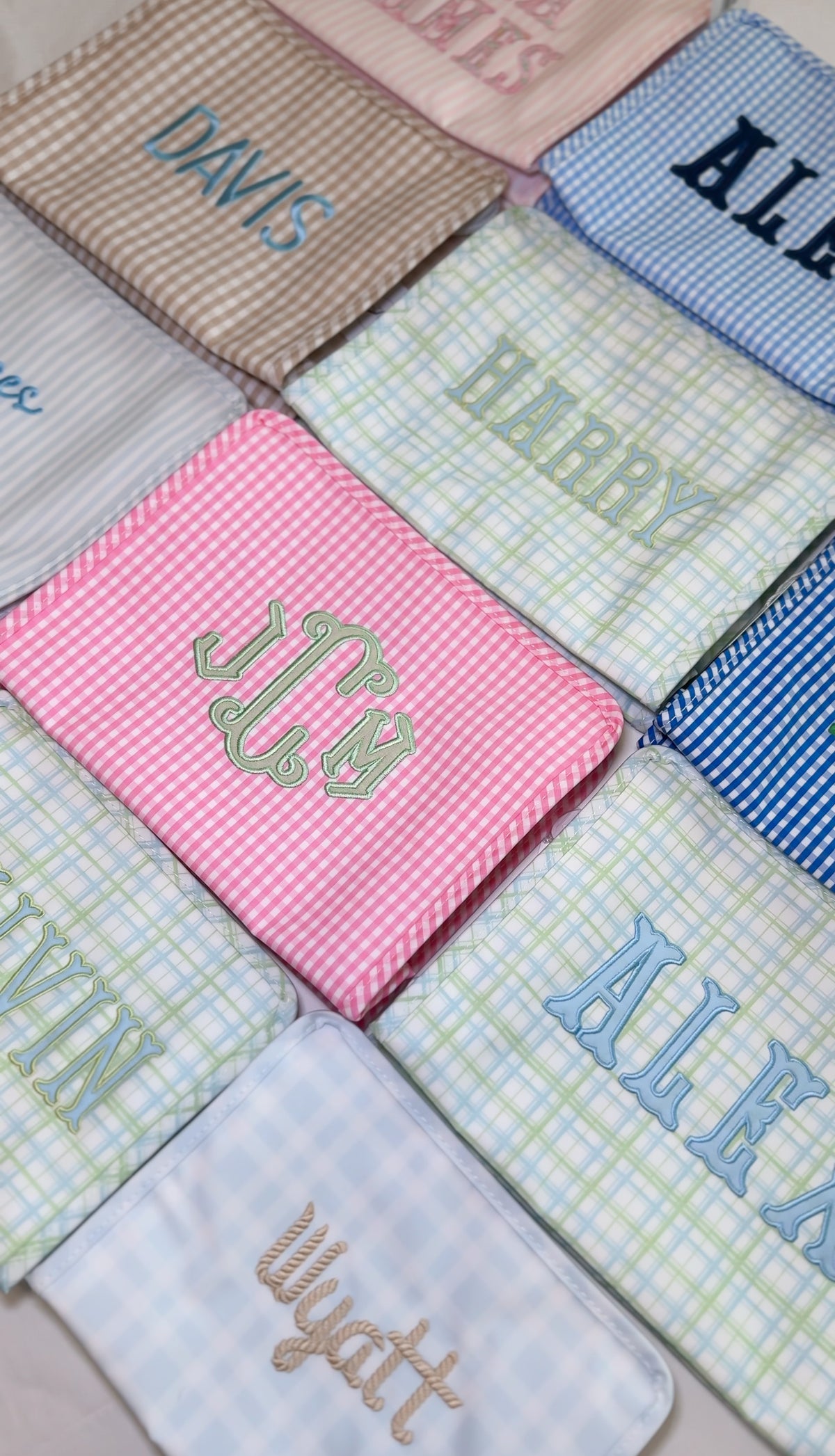 Grid of monogrammed TRVL pouches in various plaid and gingham patterns, embroidered with names including Davis, Harry, and Wyatt