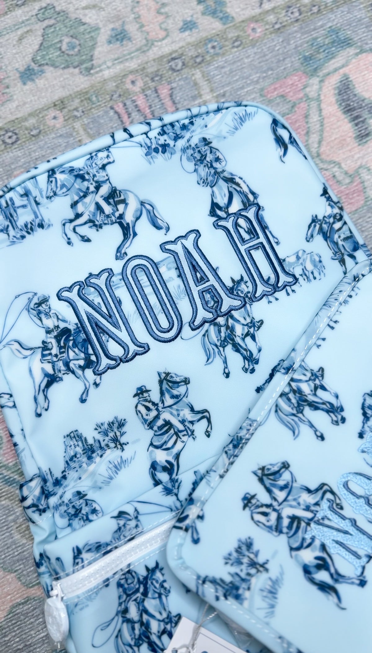 Light blue cowboy toile print TRVL Backpacker embroidered with the name 'Noah', a matching bag visible beside it