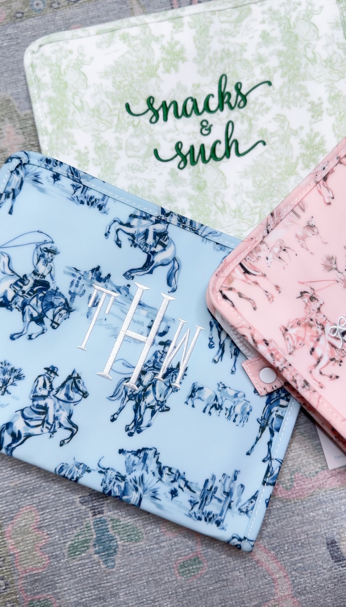 Blue and pink cowboy toile TRVL pouches with silver 'THW' monogram embroidery, next to a green toile pouch embroidered 'snacks & such'