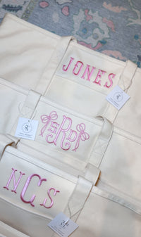 Three natural canvas TRVL Maxi Totes stacked, embroidered with 'Jones', a pink bow monogram, and an 'NCS' monogram