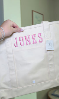 Natural canvas TRVL Maxi Tote held up, embroidered with the name 'Jones' in pink lettering and a TRVL Designs tag
