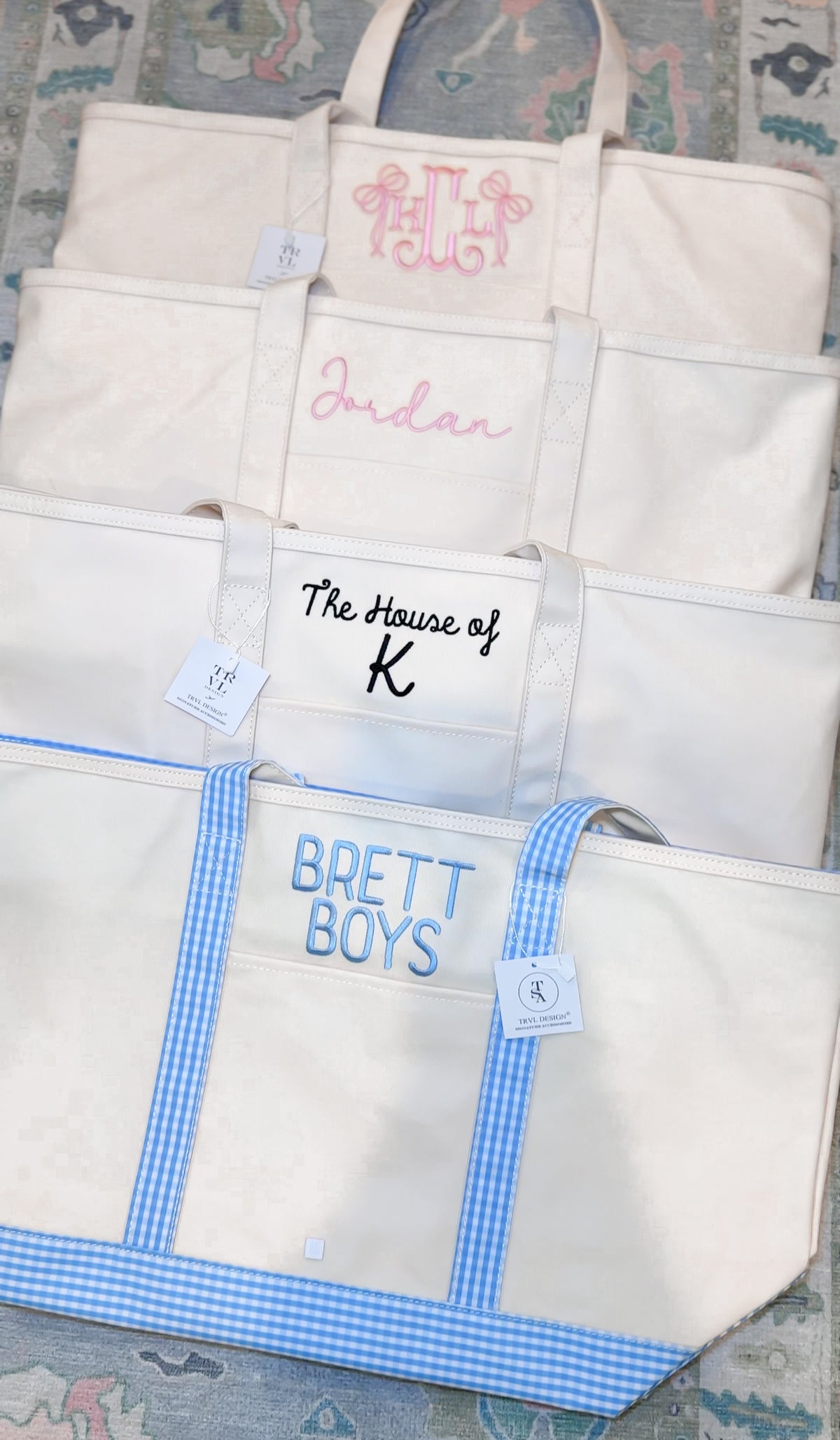 Four natural canvas TRVL Maxi Totes stacked, embroidered with a pink bow monogram, 'Jordan' in script, 'The House of K', and 'Brett Boys' in blue with gingham handles