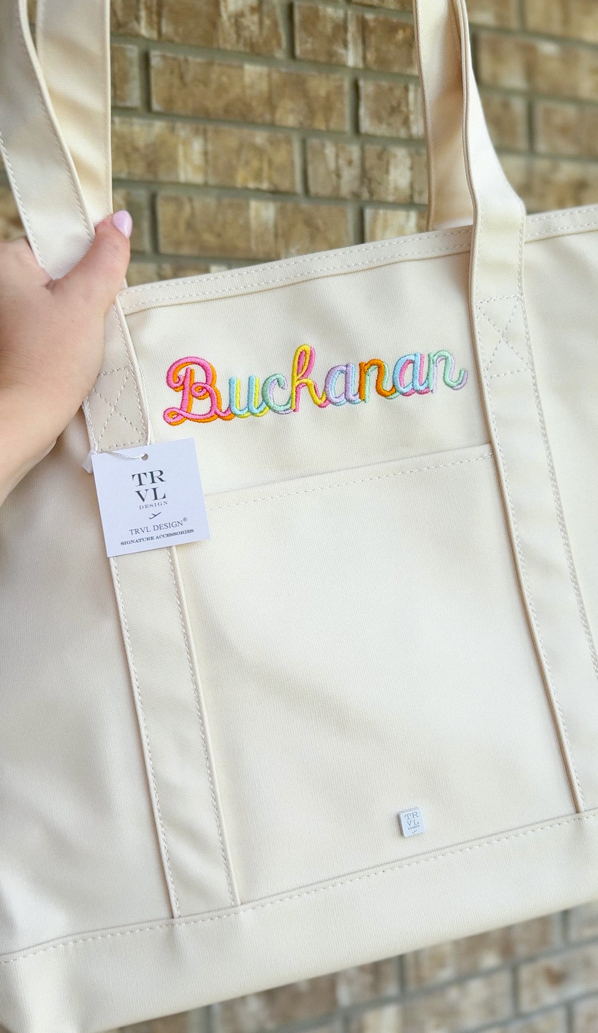 Natural canvas TRVL Maxi Tote held up, embroidered with the name 'Buchanan' in rainbow-colored script