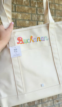 Natural canvas TRVL Maxi Tote held up, embroidered with the name 'Buchanan' in rainbow-colored script