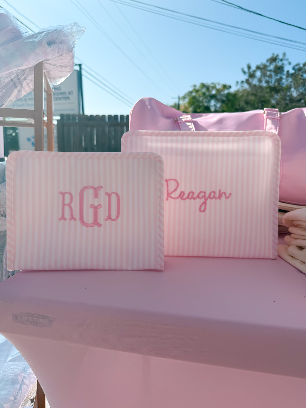 Pink Pimlico stripe TRVL Roadie pouches embroidered with 'RGD' monogram and 'Reagan' in cursive script, shown outdoors with a pink duffel bag