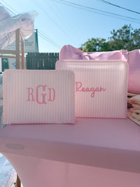 Pink Pimlico stripe TRVL Roadie pouches embroidered with 'RGD' monogram and 'Reagan' in cursive script, shown outdoors with a pink duffel bag