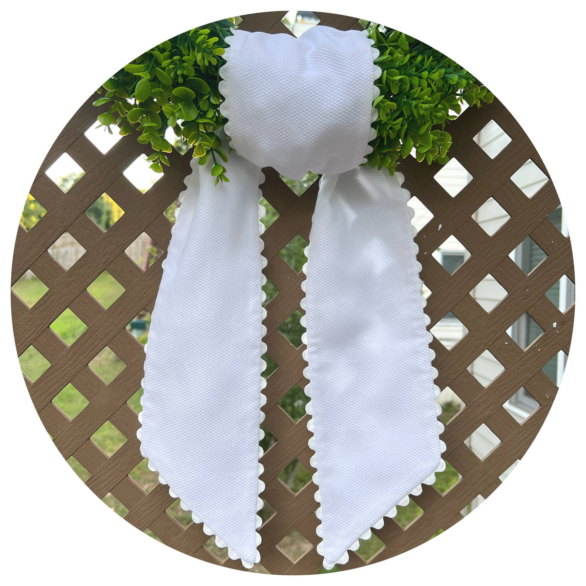 White cotton pique sash tied on a boxwood wreath, white scalloped trim, close-up view on a lattice backdrop