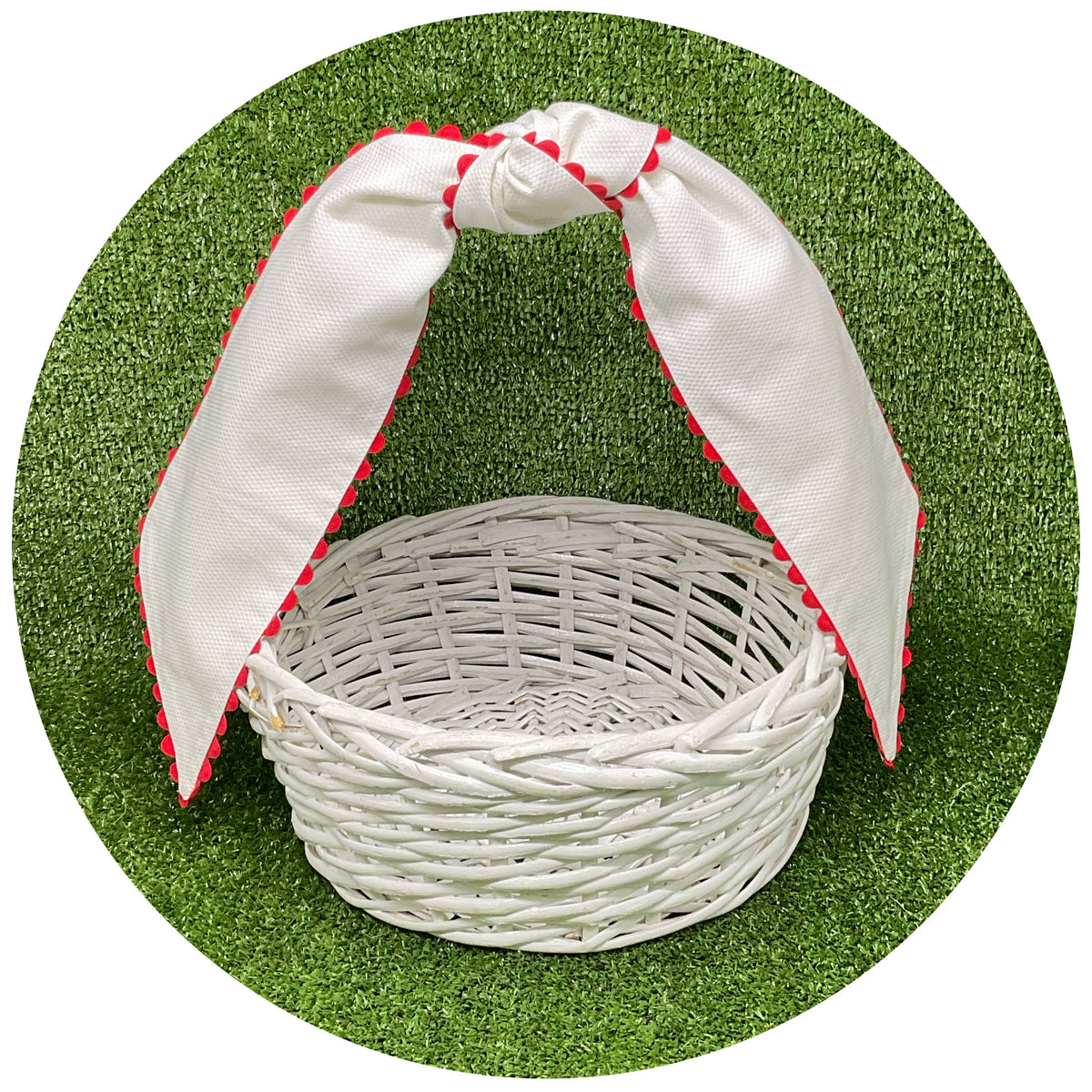 White cotton pique sash with red scalloped trim tied in a bow on a white wicker basket handle, styled against artificial turf