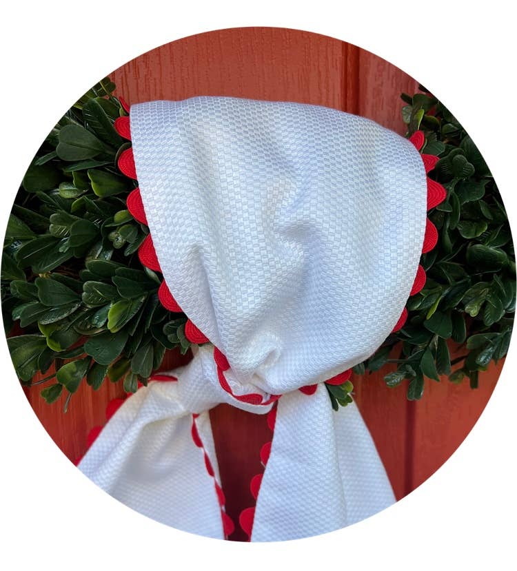 Close-up of white cotton pique sash with red scalloped trim tied on a boxwood wreath against a red wood door